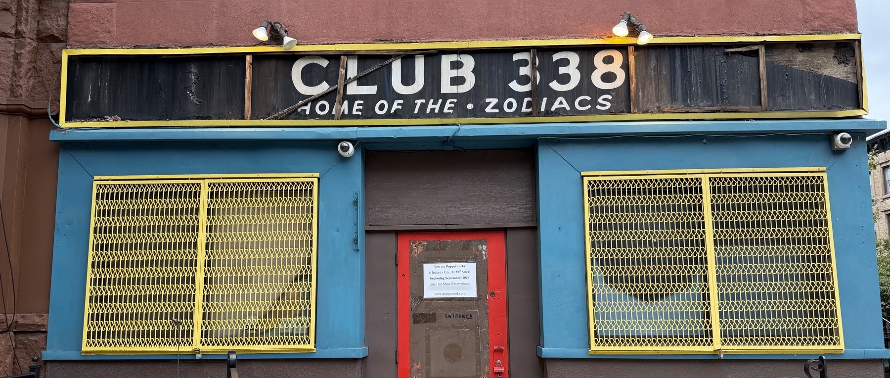 The storefront at 338 Sixth Avenue. A black board with cream letters reads CLUB 338, HOME OF THE ZODIACS, above a teal front, yellow window grilles and a red door.