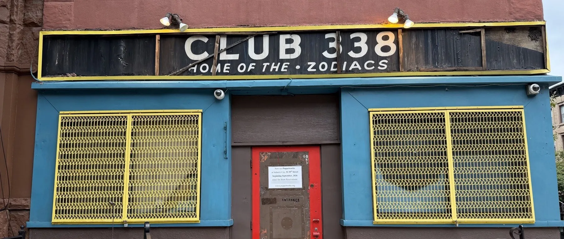 The storefront at 338 Sixth Avenue. A black board with cream letters reads CLUB 338, HOME OF THE ZODIACS, above a teal front, yellow window grilles and a red door.